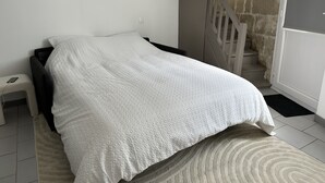 Une chambre à coucher avec un lit, une petite table de chevet, une commode, un mur blanc, un luminaire au plafond et un escalier.