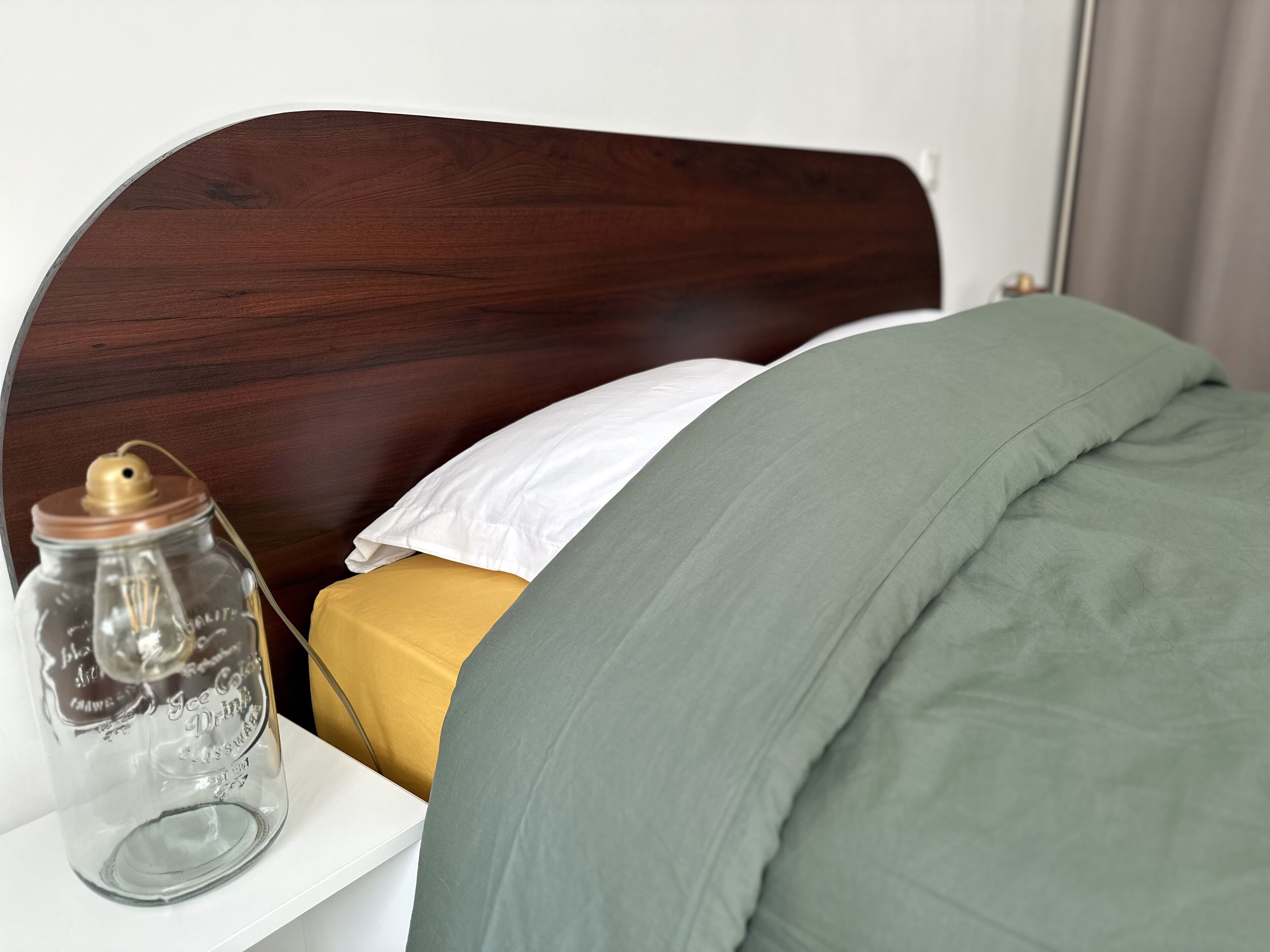 A bed with a wooden headboard, a white pillow, a mustard-colored blanket, and a green blanket.