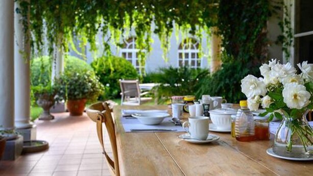 An outdoor dining area with a wooden table set for a meal, surrounded by greenery and a white building in the background. - Modern Studio set an Expansive Garden - 2253 (Harare)