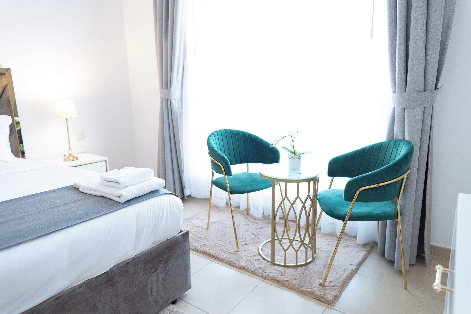 A modern hotel room with a large bed, two teal chairs, a small round table, and a potted plant.
