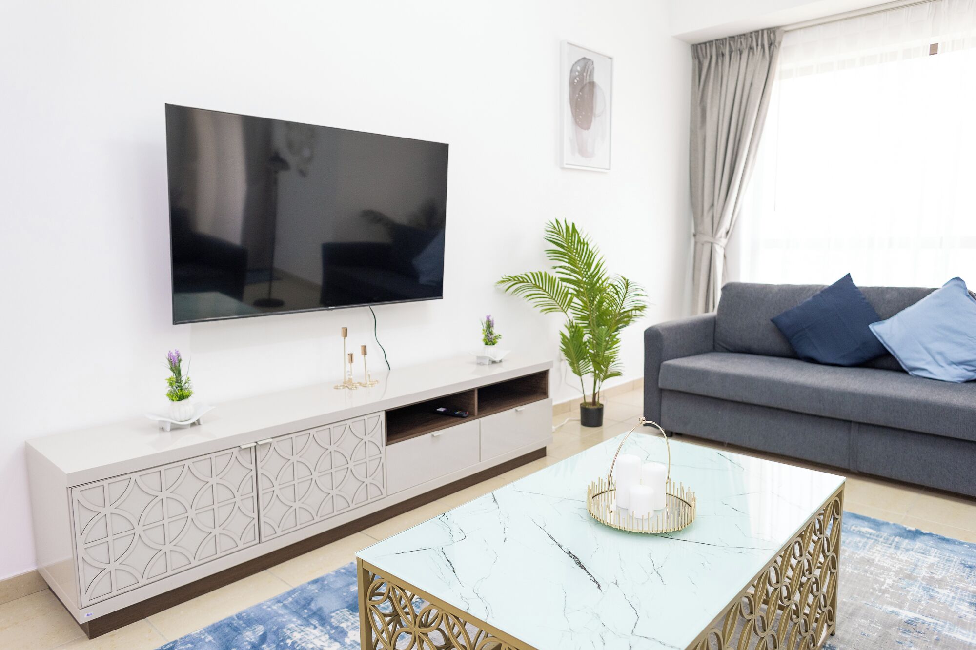 A modern living room with a flat-screen TV mounted on a white cabinet, a grey sofa with blue cushions, a marble coffee table, and a potted plant.