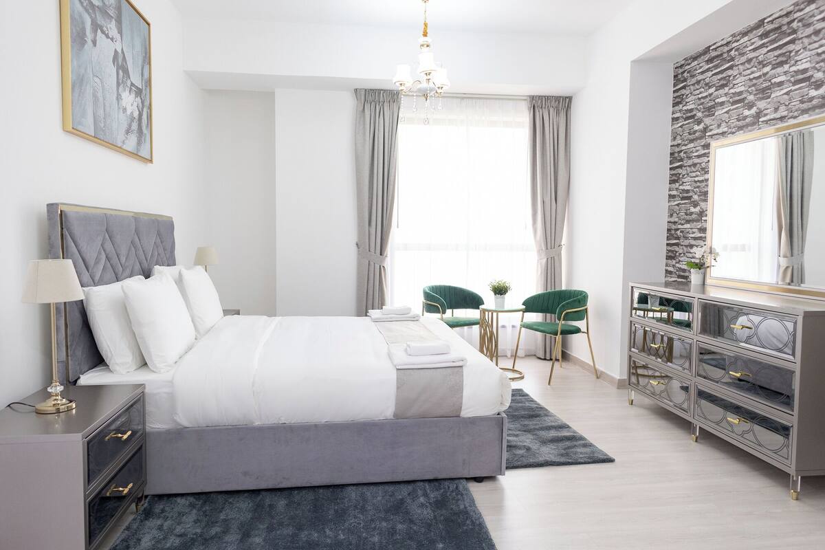 A modern bedroom with a large bed, a bedside table, a dresser, a chandelier, and a stone-textured wall.
