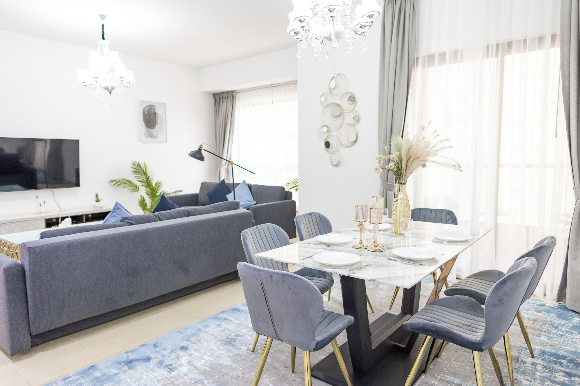 A modern living room with a grey sofa, a dining table set with white plates and gold candlesticks, and a chandelier above.