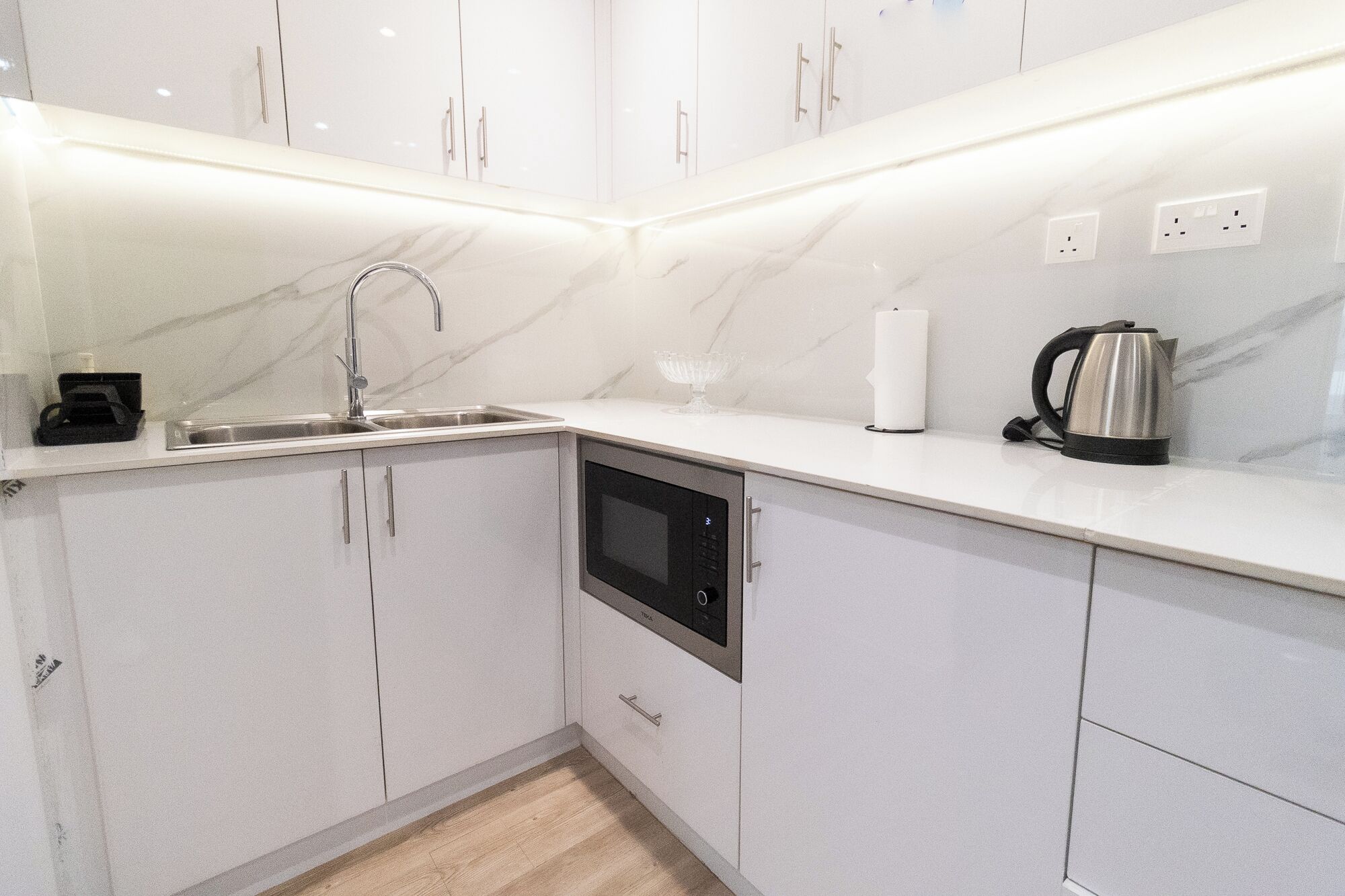 A modern kitchen with white cabinets, a built-in microwave, a kettle, and a sink.