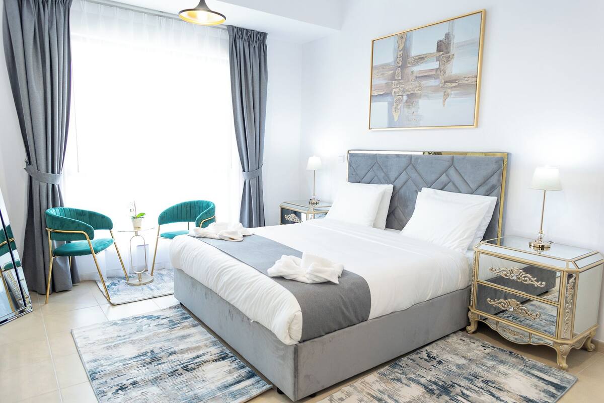 A modern bedroom with a large bed, two chairs, a bedside table with a lamp, and a painting on the wall.