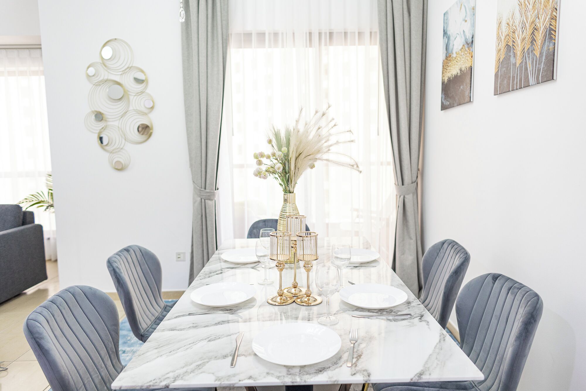 Un comedor con una mesa de mármol, sillas tapizadas en gris y un jarrón con flores secas.