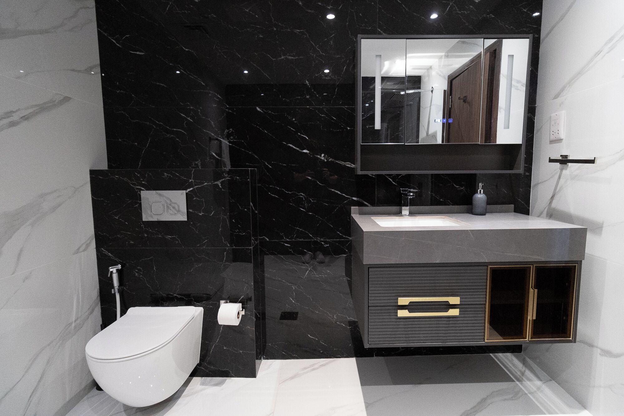 Un baño moderno con pared de mármol negro, inodoro blanco, lavabo gris y un mueble con pomo dorado.