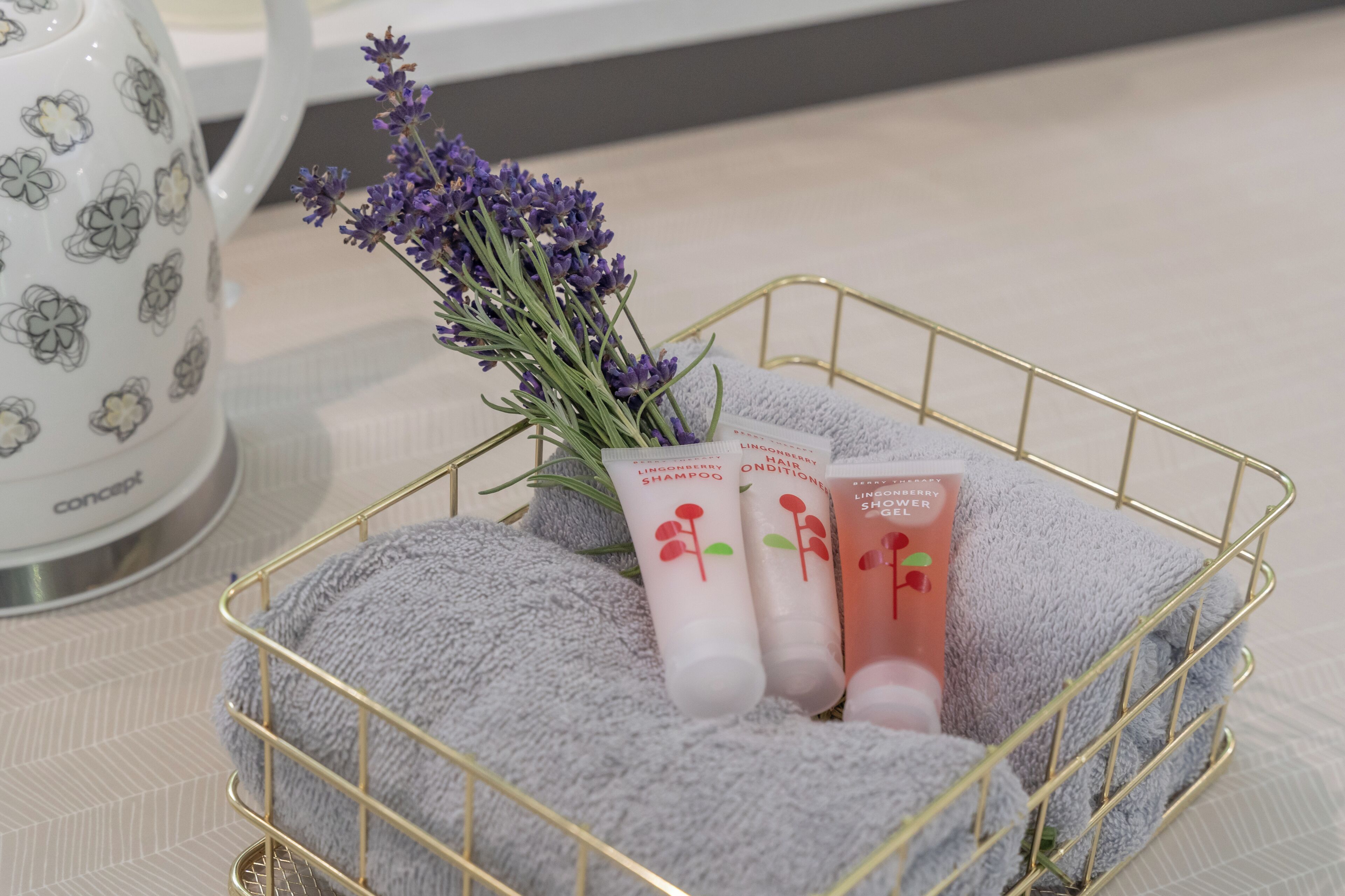 A gold wire basket containing lavender, a grey towel, and two bottles of shampoo and conditioner.