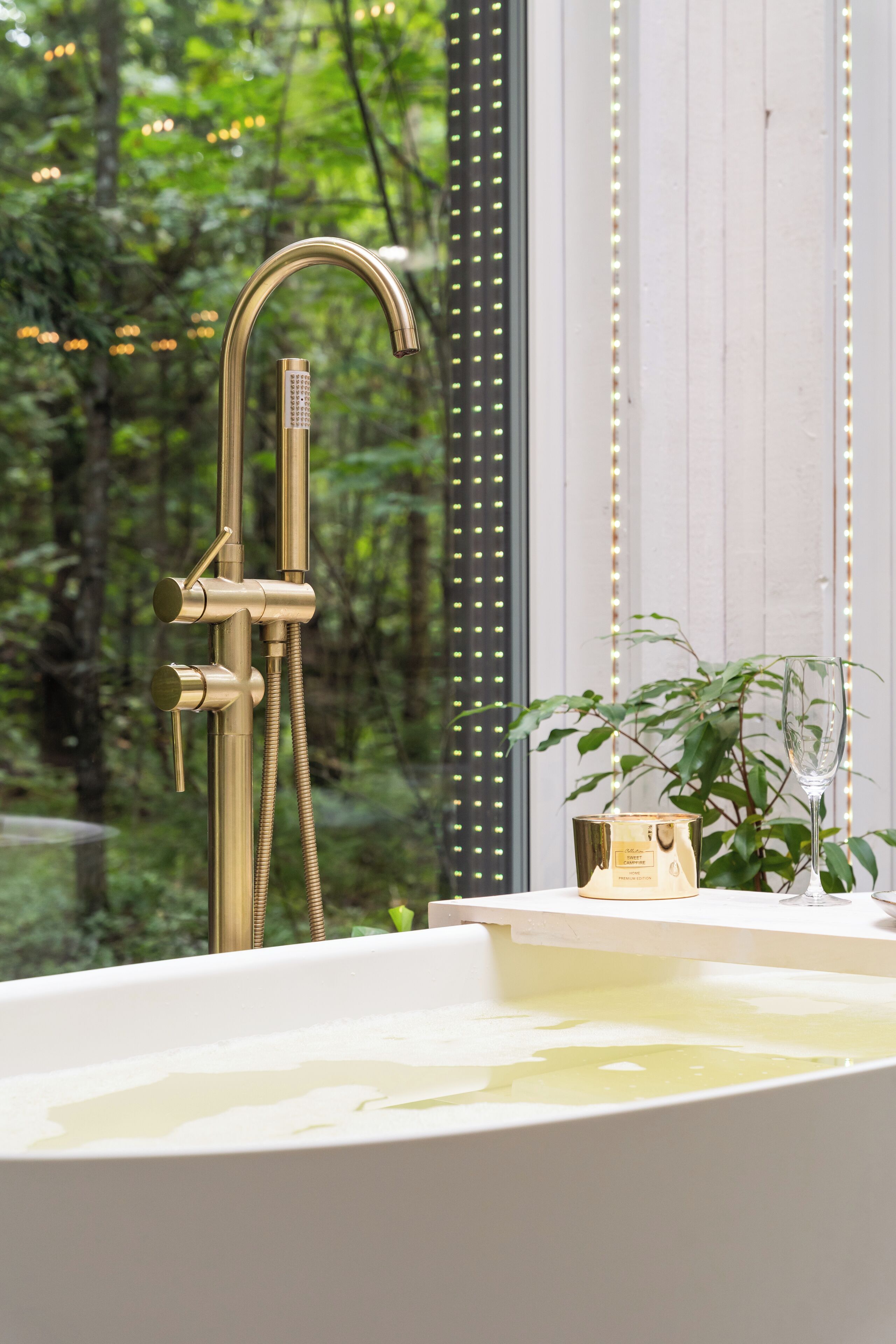 A modern bathroom with a gold showerhead, a glass bathtub, and a view of a forest through a large window.