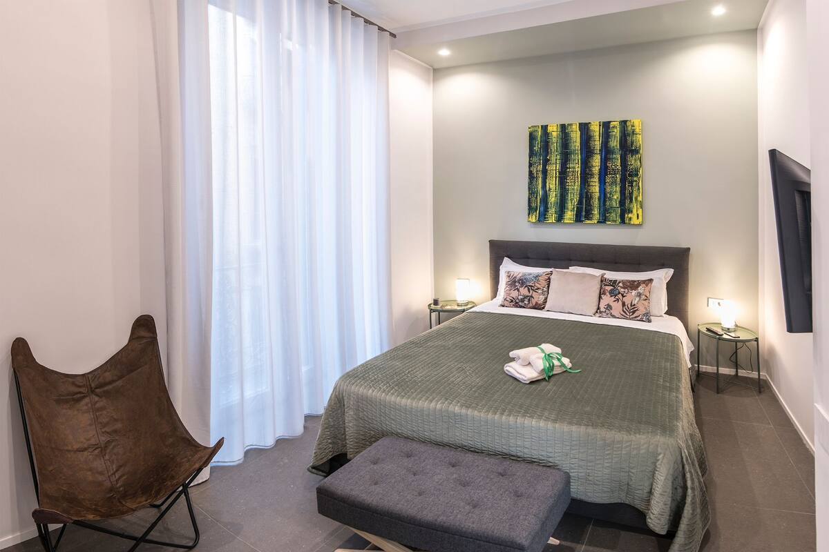 A modern bedroom with a large bed, a chair, a bench, and a painting on the wall.