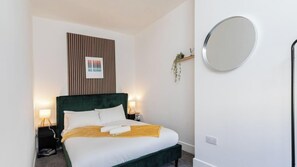 Un lit bien fait, avec une tête de lit verte, deux lampes de chevet, un miroir fixé au mur et un panneau de bois vertical orné d’une citation inspirante.