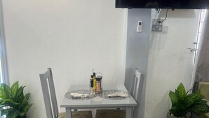 Un comedor con una mesa dispuesta para dos personas, dos sillas, un televisor montado en la pared y plantas en macetas a ambos lados.