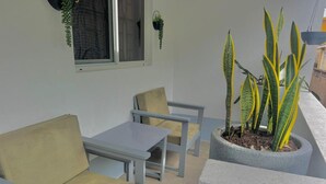 Un pequeño espacio al aire libre con dos sillas y una mesa, una planta en maceta con hojas amarillas y verdes, y una ventana con cortina.