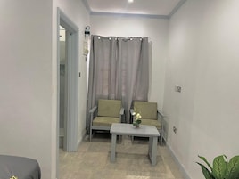 Um quarto pequeno e estreito com dois sofás e uma mesa pequena, uma porta que dá para outro espaço, e uma planta no canto.