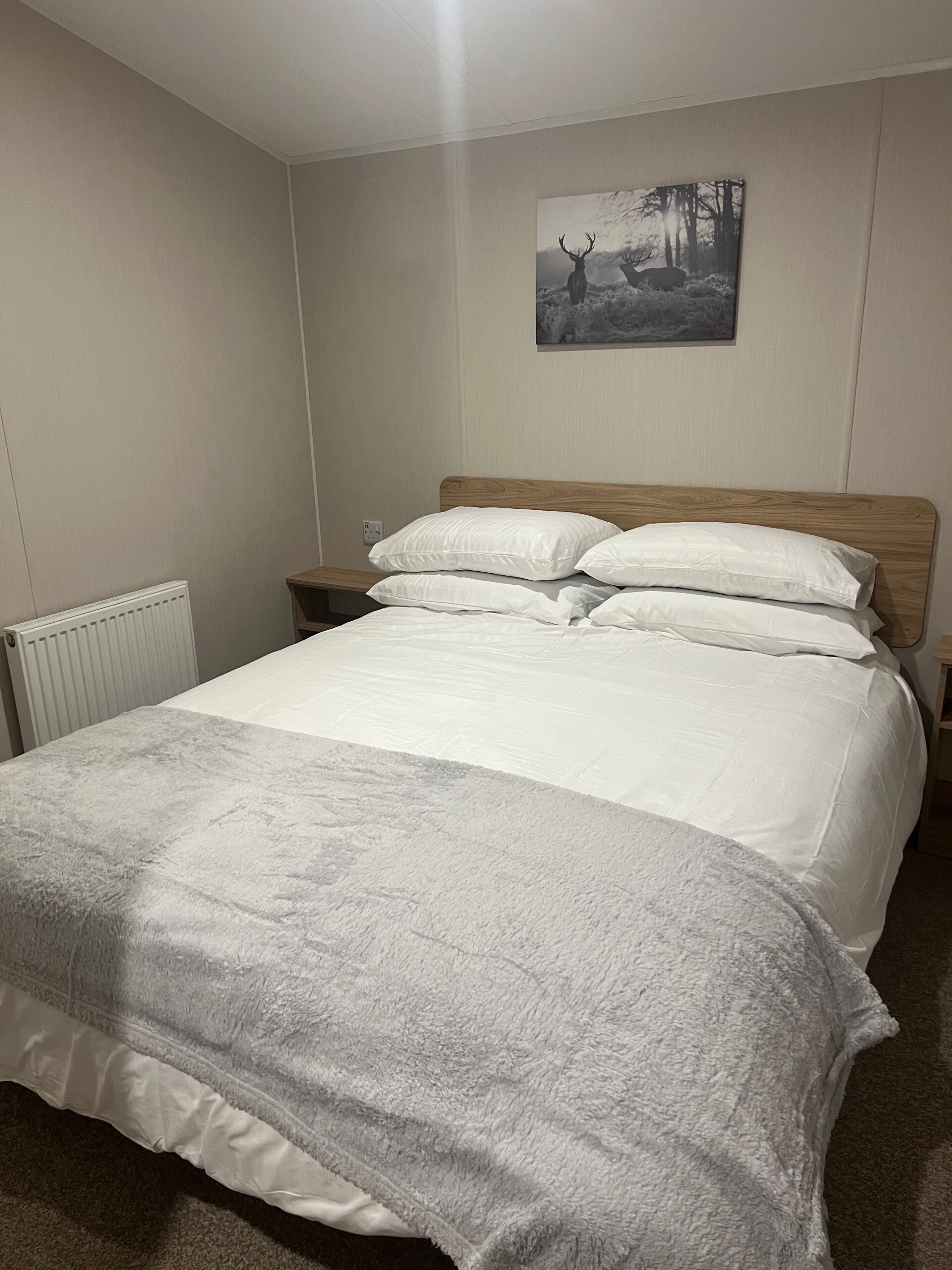 Un lit bien fait, avec du linge de lit blanc et une couverture grise, une tête de lit en bois et une image de cerfs accrochée au mur.