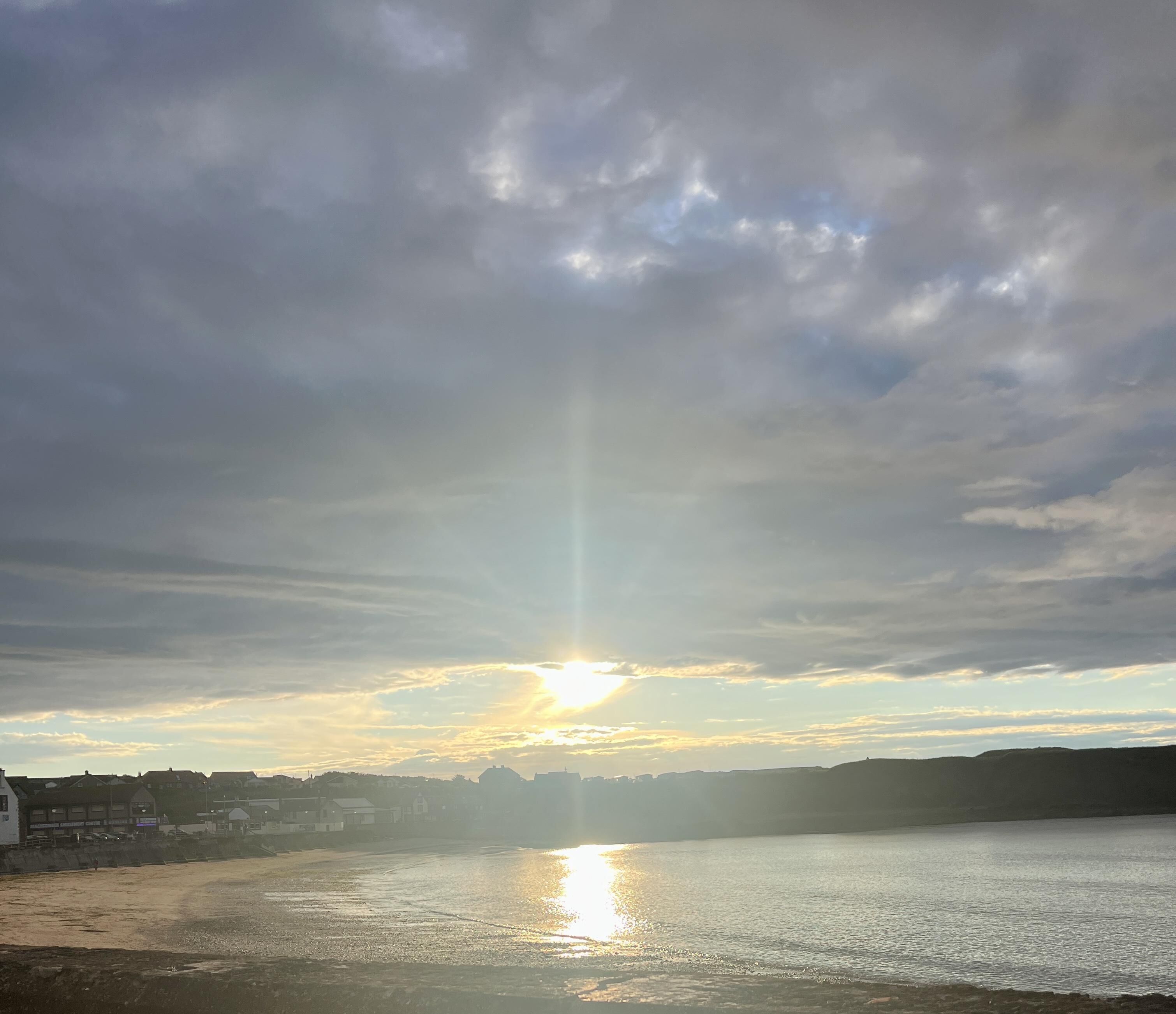 A coastal scene with the sun setting over a body of water, casting a reflection on the surface.