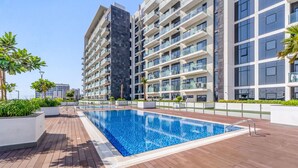 Un moderno edificio residencial con piscina en la azotea, rodeado de vegetación y con balcones con barandillas de vidrio.