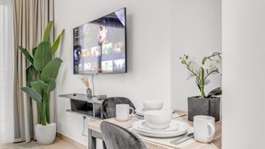 Un área de comedor moderna con un televisor en la pared mostrando un menú, una mesa de comedor con vajilla blanca y una planta.