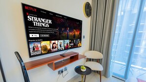 A modern living room with a large flat-screen TV displaying a Netflix series. There is a wooden shelf below the TV, a small round table with a plant, and a comfortable chair. - Chic Studio Oasis in Dubai (Dubai)