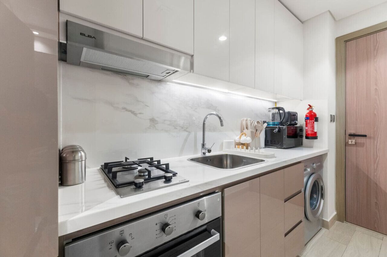 A modern kitchen with a built-in stove, sink, and washing machine, featuring white cabinetry and a marble countertop.