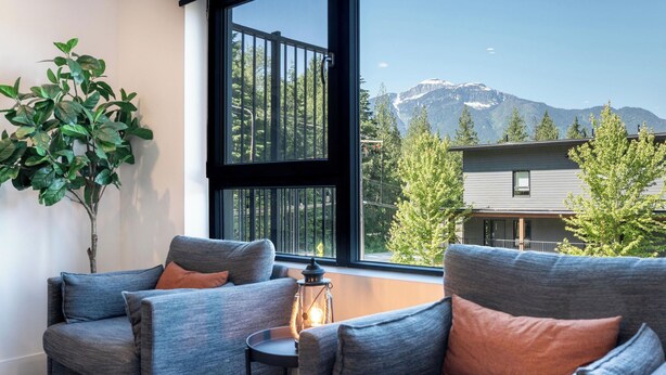 A room with two grey sofas, a small table, a potted plant, and a view of mountains through a large window. - Cedar and Loam by Revelstoke Vacations (Revelstoke)
