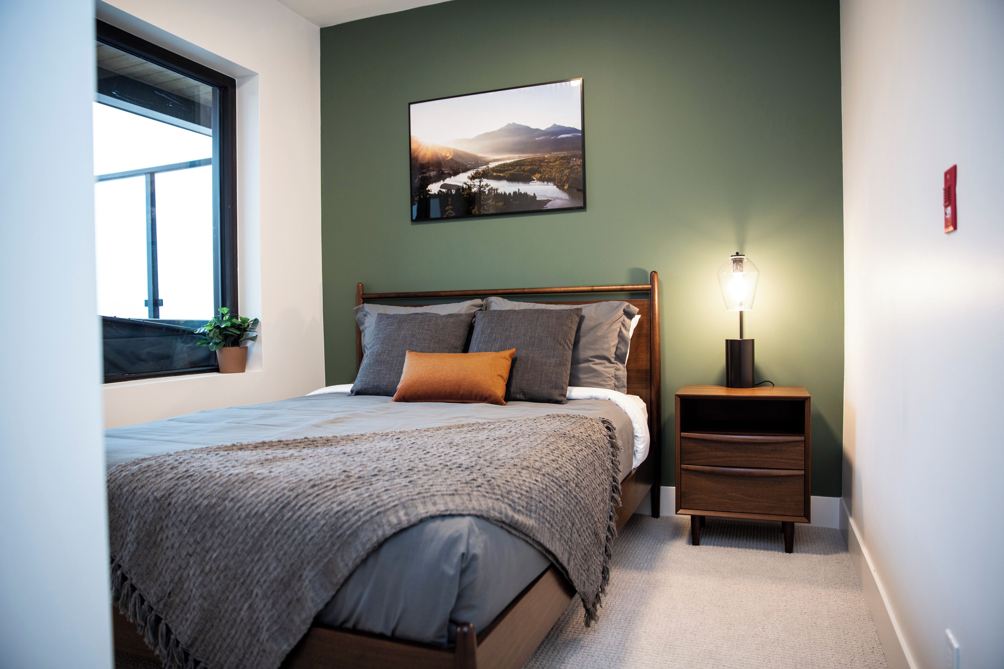 A bedroom with a bed, a nightstand, a lamp, a window, and a wall-mounted picture.