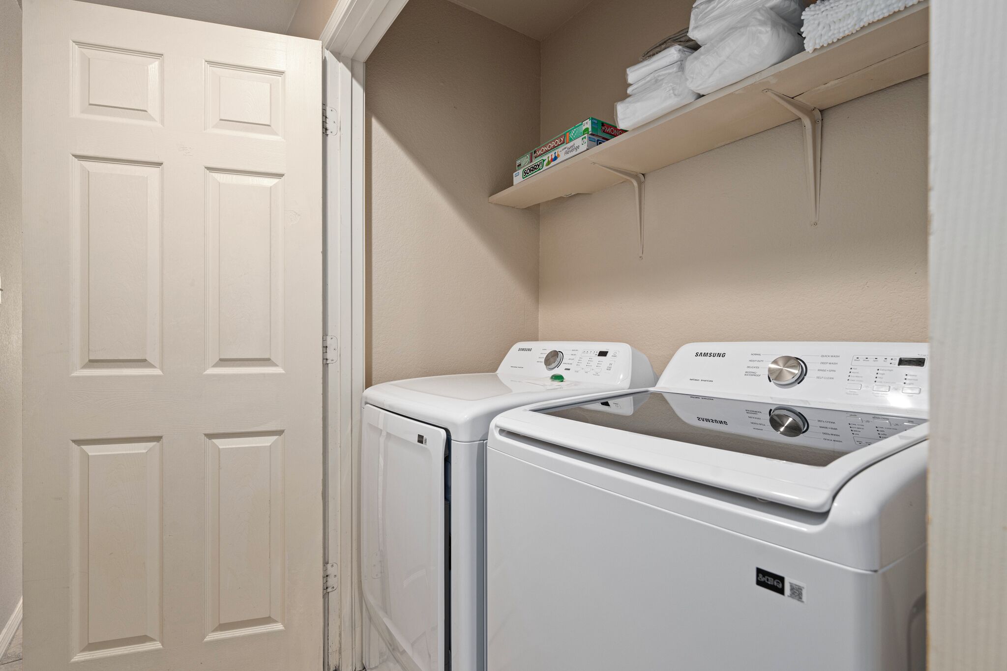 A laundry room with two front-loading washers and dryers, a white door, and a shelf with folded towels.
