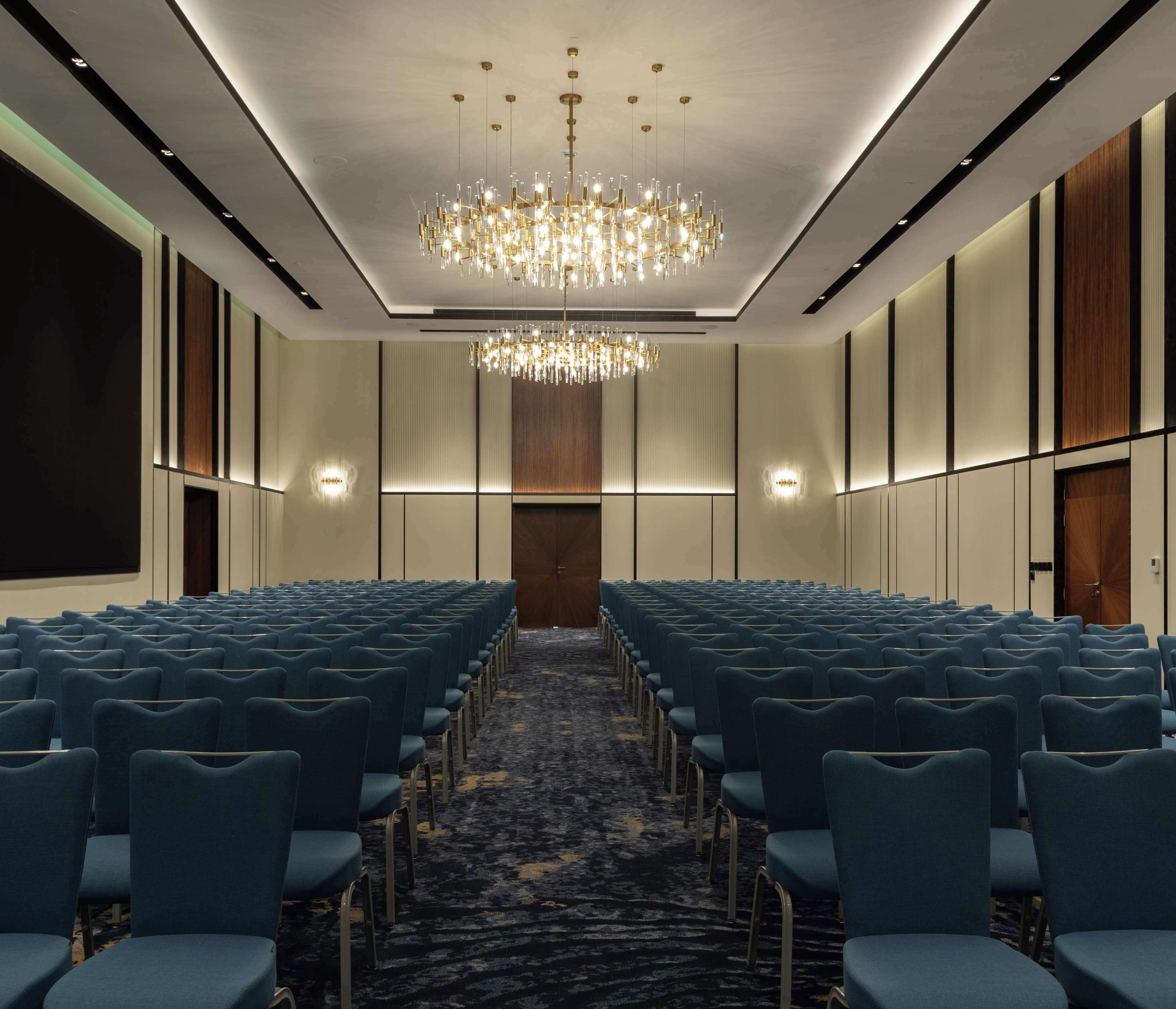 A conference room with rows of blue chairs, a patterned carpet, a chandelier, and wall-mounted lights.