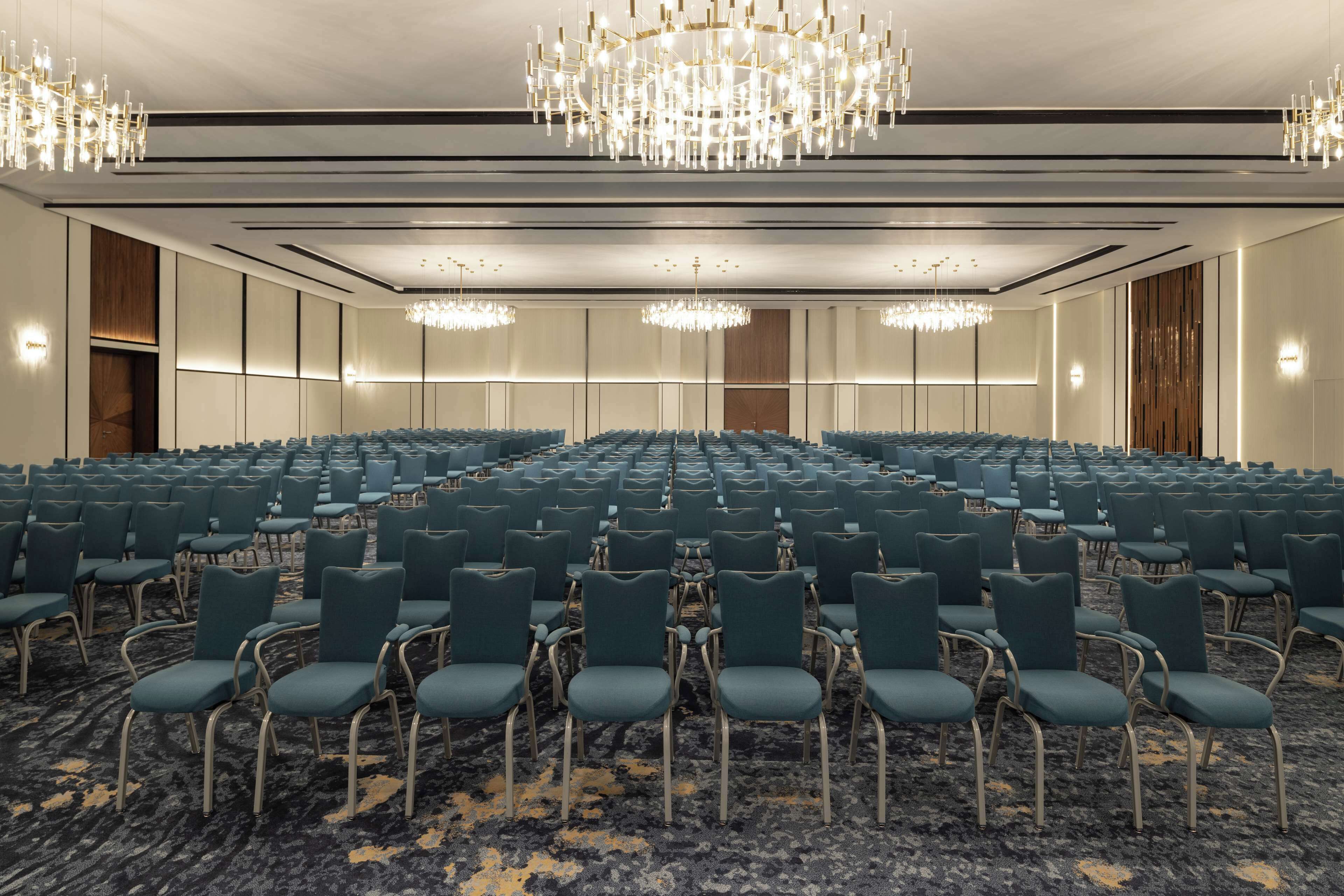 Un salón de conferencias con hileras de sillas color turquesa, una alfombra estampada y candelabros.