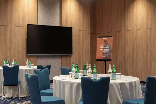 A conference room with round tables, blue chairs, and a large flat-screen TV on the wall. - Park Inn By Radisson Hotel & Apartments Kuwait (Mahboula)