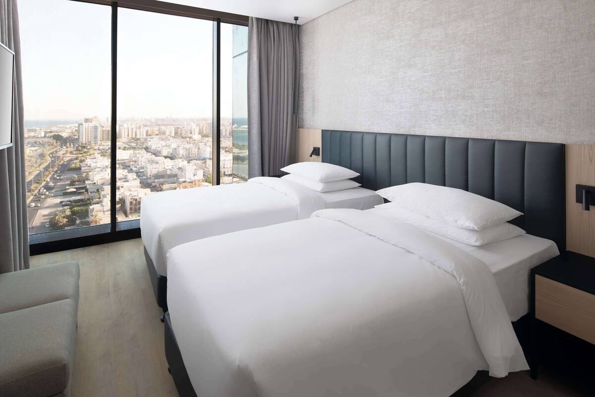 A hotel room with two beds, a large window with a city view, a modern headboard, and a bedside table.