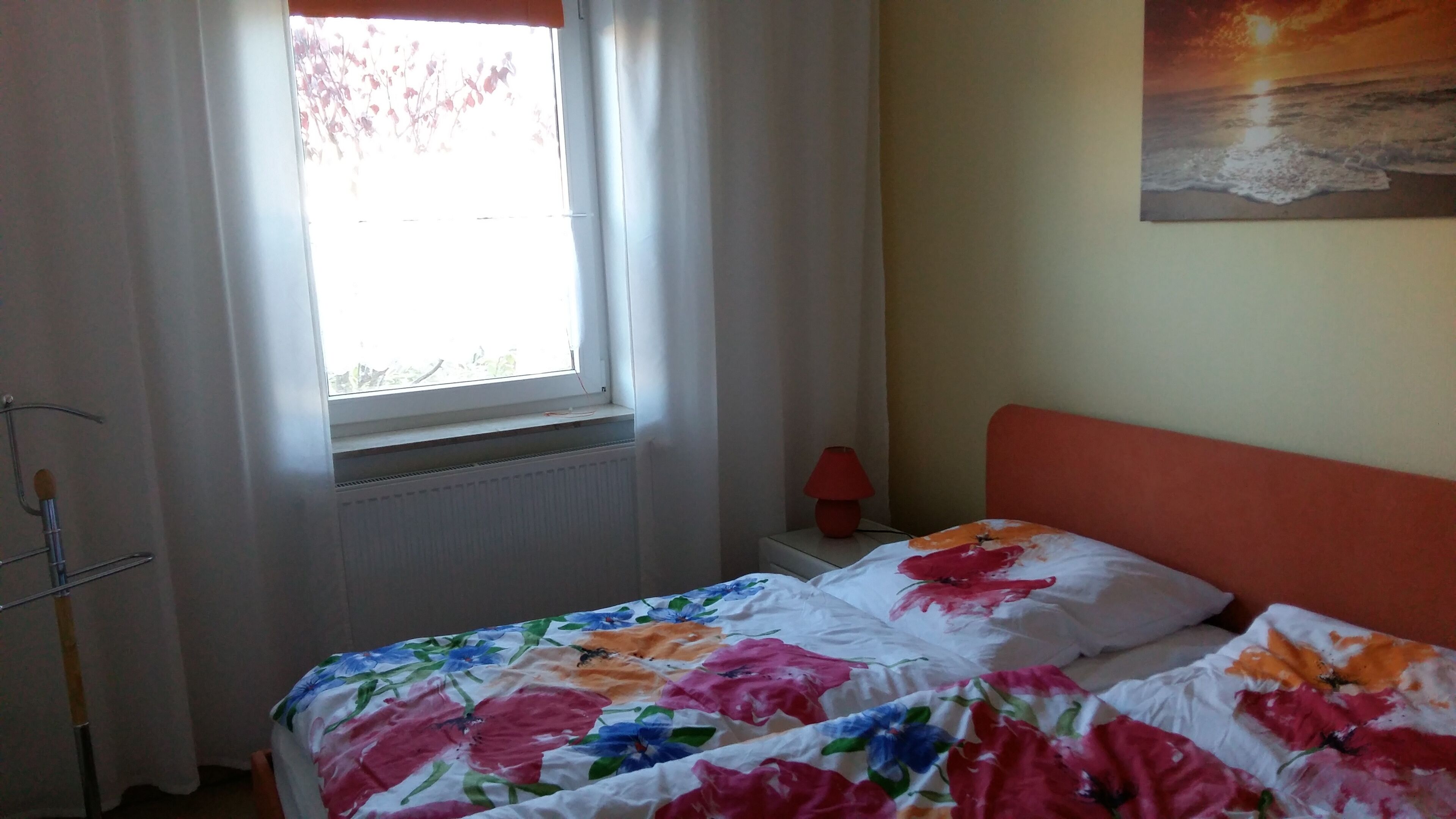 Een slaapkamer met een bed, een raam met gordijnen, een radiator, een nachtkastje met een lamp en een schilderij aan de muur.