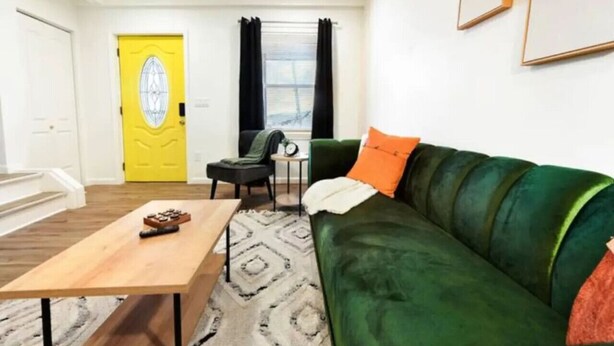 A living room with a green sofa, a wooden coffee table, a yellow door, and a window with curtains. - 3 BedHome in Upper Lawrenceville (Pittsburgh)