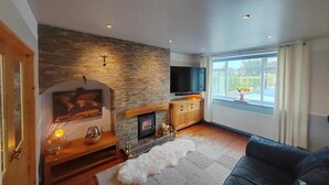 A living room with a stone fireplace, a flat-screen TV mounted above it, a sofa, and a large window with curtains. - Cozy Blackpool Retreat With 3 Bedrooms (Blackpool)