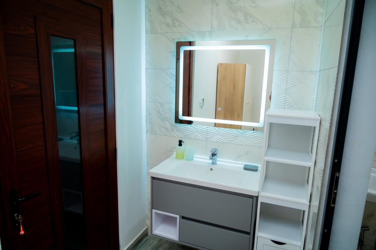 A bathroom with a white vanity, a mirror with integrated lighting, and a wooden door.