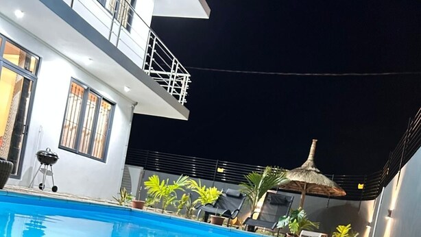 A swimming pool with a concrete edge and a stone border, a covered outdoor area with a thatched roof, and a building with a balcony and railing. - Villa boulevard Paradise ideal for relaxing at will with family or friends (Grand Baie)