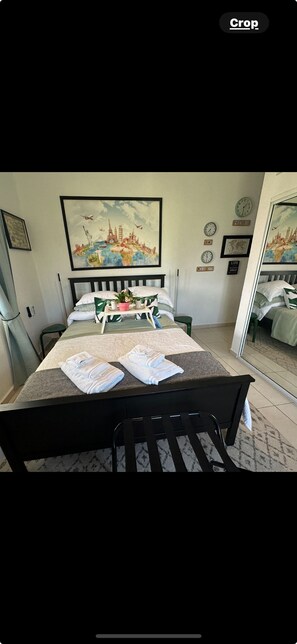 A neatly made bed with pillows and a tray of towels, a painting of a cityscape on the wall, and a mirror reflecting the room.