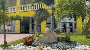 A yellow building with a flag, surrounded by a well-maintained garden with rocks and plants. - Sunny Villa Mar Chiquita, WE HAVE WATER  & Game room-beach hopping paradise (Tierras Nuevas Poniente)