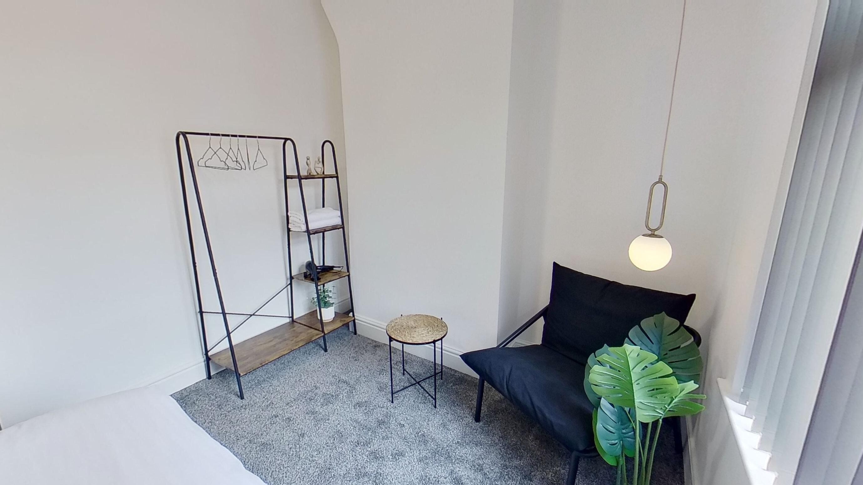 A room with a black chair, a round table, a metal shelf unit, and a hanging light fixture.