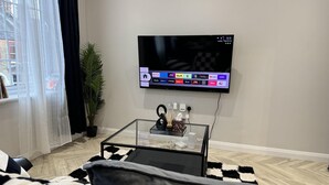 Un salon moderne avec un téléviseur à écran plat fixé au mur, une table basse en verre agrémentée d’objets décoratifs, un tapis à carreaux noirs et blancs et une grande plante en pot.