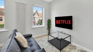 Un salon moderne avec un canapé en cuir noir, une table basse en verre et un téléviseur fixé au mur qui affiche le logo « NETFLIX ».
