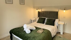 Un lit bien fait, recouvert d’une couverture verte, avec deux oreillers et une serviette enroulée. Deux lampes murales sont fixées de part et d’autre du lit.