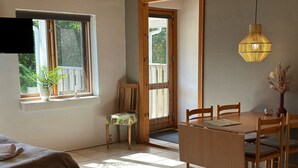 Un coin repas avec une table et des chaises en bois, une petite table d’appoint sur laquelle est posé un vase, et une fenêtre donnant sur un paysage verdoyant.