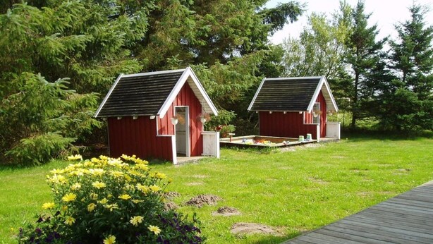 Deux cabanes en bois rouge, dotées de toits inclinés, entourées d’une zone herbeuse et d’une forêt dense.