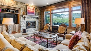A cozy living room with a stone fireplace, comfortable seating, and large windows offering a scenic view. - Gorgeous Deer Valley Home Minutes From the Slopes (Park City)