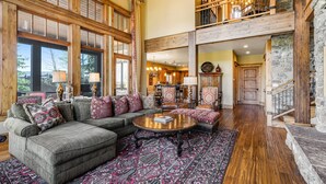 A spacious living area with a large sectional sofa, a round coffee table, and a patterned rug. There is a staircase leading to an upper level with a balcony. - Gorgeous Deer Valley Home Minutes From the Slopes (Park City)