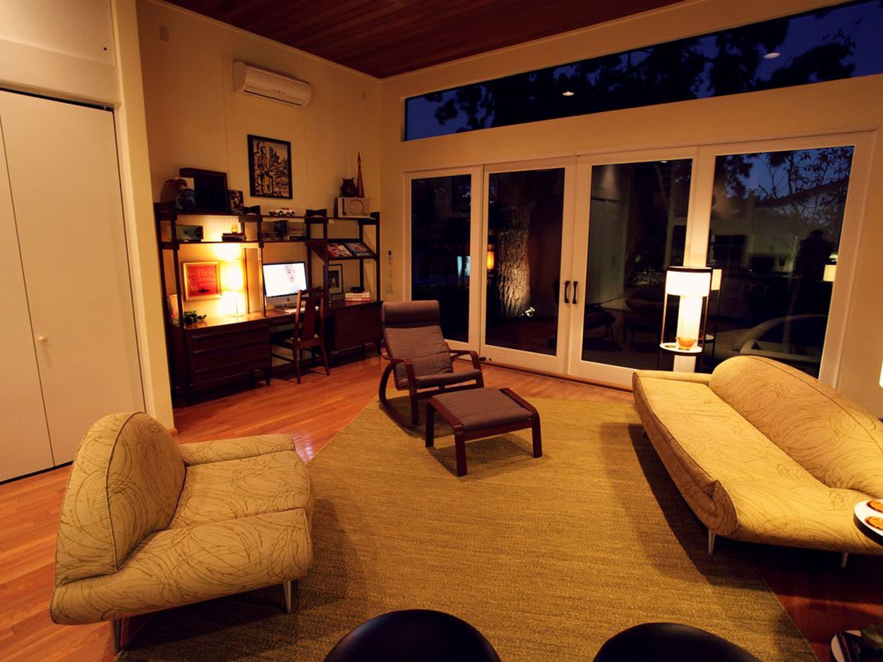 A living room with a sofa, armchairs, a coffee table, and a desk with a computer. There is a bookshelf and a window with a view of trees.