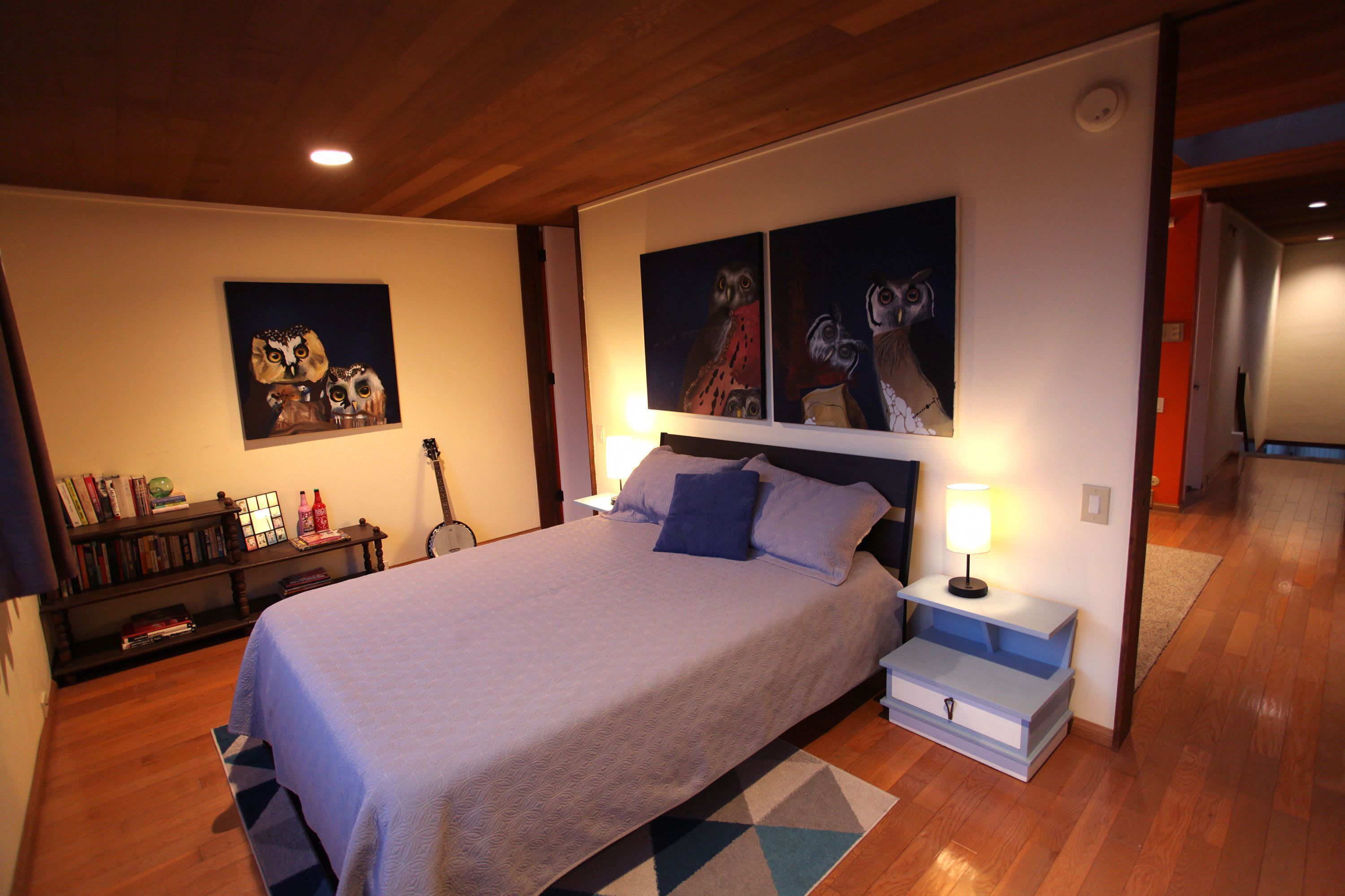 Un dormitorio con una cama grande, una mesita de noche con lámpara, una estantería y dos cuadros de búhos colgados en la pared.