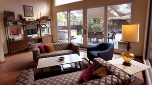 Un salon comprenant un canapé, un fauteuil et une table basse. On y trouve également une bibliothèque remplie de livres et un téléviseur. La pièce est dotée de grandes fenêtres offrant une vue sur l’extérieur.