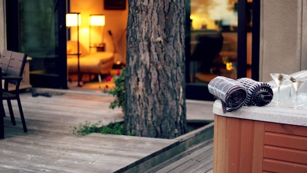 Une terrasse en bois avec un jacuzzi, un arbre et un intérieur chaleureux avec une lampe et un fauteuil.